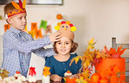 Kids celebrating Thanksgiving day. Boy in native American hat helps girl to put on head paper turkey hat.の写真素材