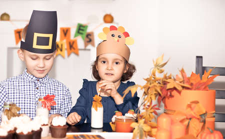 Kids sitting on festive table and celebrating Thanksgiving day. Children in paper turkey hat and pilgrim hats eating cupcakes and drinking milk.の写真素材