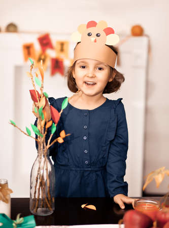 Little cute girl in paper turkey hat glues paper leaves on branches for celebrating Thanksgiving day. Diy craft art project.の写真素材