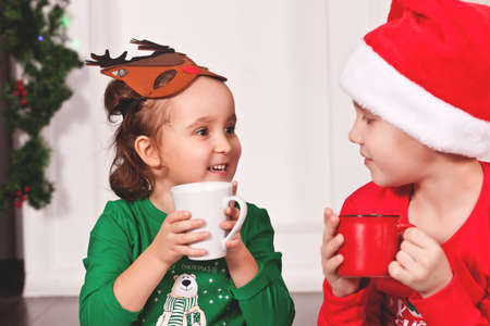 Little girl in funny deer mask and boy kid in Santa cap and Christmas pajamas drinking cocoaの写真素材
