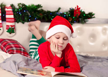Boy kid in Santa cap and Christmas pajamas reading a book.