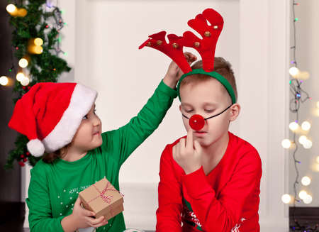 Happy little smiling girl and boy in Santa cap and deer mask sitting on the blanket in Christmas pajamas with gift box.の写真素材