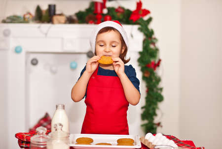 Little cute girl in red apron baking Christmas cookies at home and tastes baked cookiesの写真素材