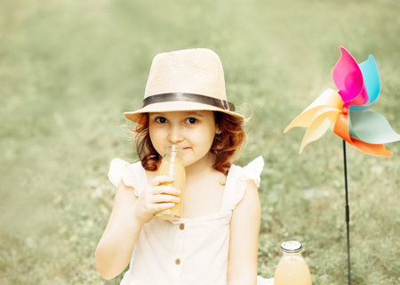 Cute little girl in a hat sitting on a blanket on a park meadow holding a bottle and drinking orange juice. Picnic on nature.の写真素材
