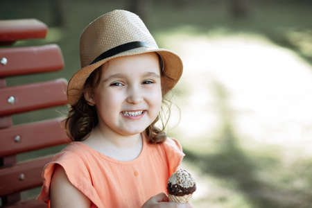 Cute little kid girl in a hat eating ice cream cone. Child sitting on a park bench and laughing ....