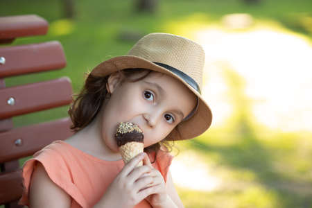 Cute little kid girl in a hat eating ice cream cone. Child sitting on a park bench. Summer concept.の写真素材