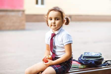 little cute schoolgirl in uniform sitting on a bench and eating an appleの写真素材