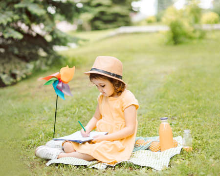 Cute little girl in a hat sitting on a blanket and writing or drawning at a picnic in natureの写真素材