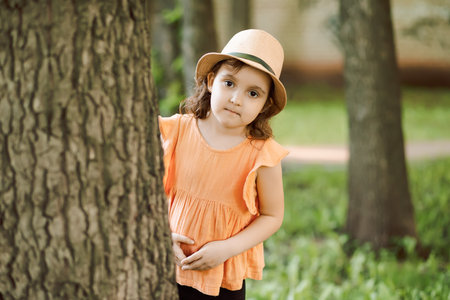 Little cute girl kid in a hat looking out from behind a tree. summer activity. happy childhoodの写真素材