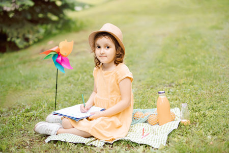 Cute happy little girl in a hat having fun in park meadow. Child sitting on a blanket and looking at camera. Picnic on nature.の写真素材