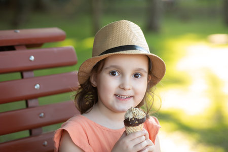 Cute little kid girl in a hat eating ice cream cone. Child sitting on a park bench and laughing. summer conceptの写真素材