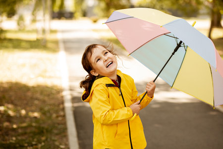 Little cute girl kid child in a yellow raincoat with multicolored rainbow umbrella laughs and looks up outdoorsの写真素材