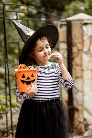 Little cute girl in witch costume holding jack-o-lantern pumpkin bucket with candies and sweets. Kid trick or treating in Halloween holidayの写真素材