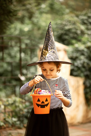 Little cute girl in witch costume holding jack-o-lantern pumpkin bucket with candies and sweets. Kid trick or treating in Halloween holidayの写真素材