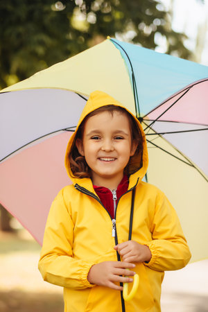 Little cute girl kid child in a yellow raincoat with multicolored rainbow umbrella laughs and looks at cameraの写真素材