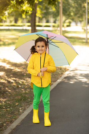 Little cute girl kid child in a yellow raincoat with multicolored rainbow umbrella laughs and looks at cameraの写真素材