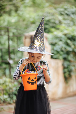 Little cute girl in witch costume holding jack-o-lantern pumpkin bucket with candies and sweets. Kid trick or treating in Halloween holidayの写真素材