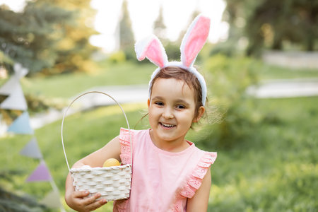 Little cute girl wear bunny ears holding basket with colorful painted eggs on Easter egg hunt in park.の写真素材