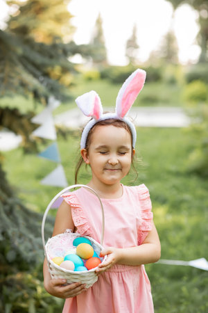 Little cute girl wear bunny ears holding basket with colorful painted eggs on Easter egg hunt in parkの写真素材
