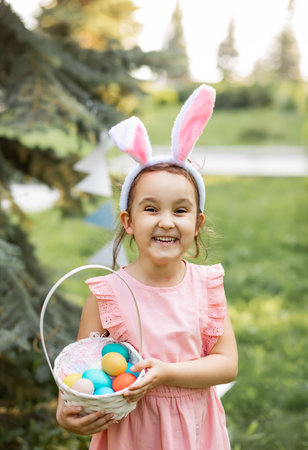 Little cute girl wear bunny ears holding basket with colorful painted eggs on Easter egg hunt in parkの写真素材