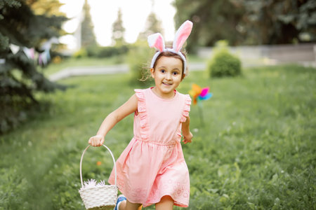 Little cute girl wear bunny ears holding basket with colorful painted eggs on Easter egg hunt in park and runningの写真素材