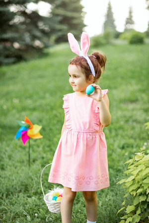 Little cute girl wear bunny ears holding basket with colorful painted eggs on Easter egg hunt in parkの写真素材
