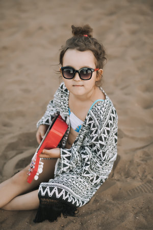 Picnic on the summer beach. Funny smiling cute little girl kid sitting on a sand and playing ukulele guitar. Happy vacation concept.の写真素材