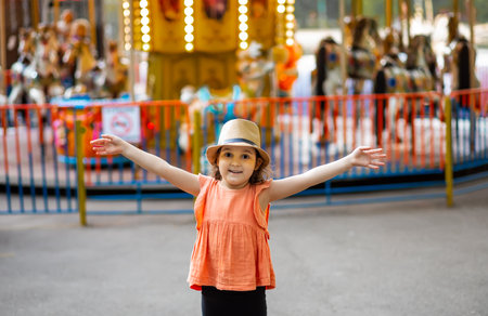Adorable little girl near the carousel outdoors in park. Happy kid in a hat standing with hands to the side. Family weekend or vacations. Summer concept.の写真素材