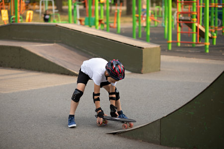 Cute kid boy child in a helmet sitting in a special area in skatepark and standing on skateboard. Summer sport activity concept. Happy childhood.の写真素材