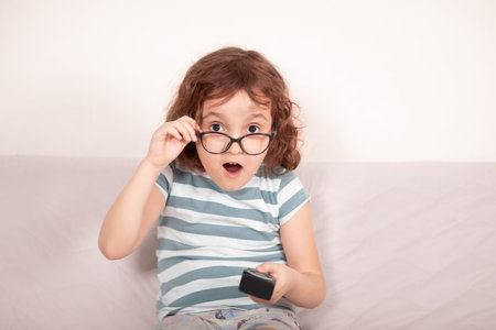 Cute curly little girl kid child in a eyeglasses surprised watching tv at home with remote control in a handsの写真素材