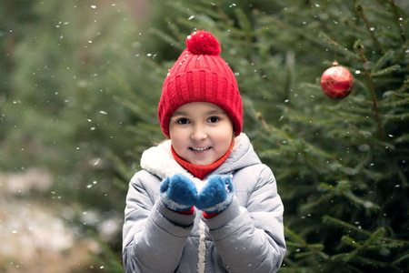 Happy kid girl in red cap playing outdoor in snowy winter forest and blowing snow. Active winter holidays celebrate concept. Happy childhoodの写真素材