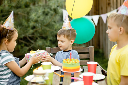 Cute funny four year old boy celebrating his birthday with family or friends with homemade baked cake in a backyard. Birthday party for kids. His gets presents gift box.の写真素材