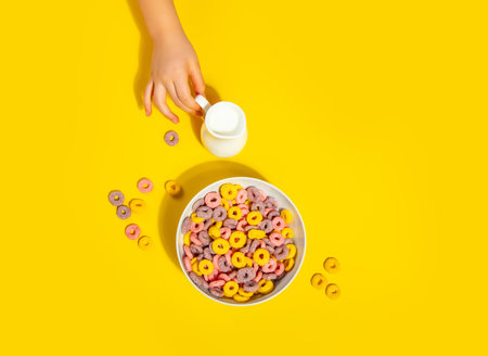 Bowl of colorful cereal corn rings on yellow table. Breakfast concept. Top view. Kid hand pouring milk into bowl.の写真素材