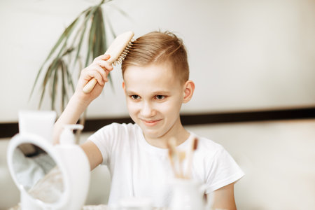 Smiling blonde teenager kid boy brushing hair with comb at home looking in small mirror. Morning routineの写真素材