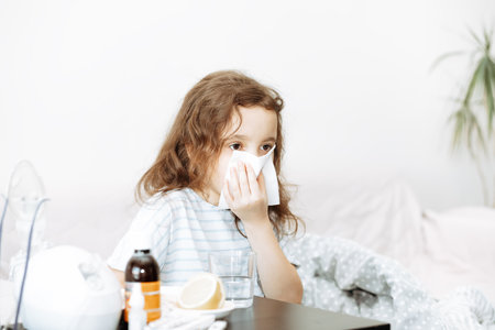 Sick little kid girl sits on bed at home and blows her nose. Flu season. Healthcare conceptの写真素材