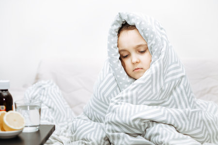 Sick sad little kid girl sits on bed at home wrapped in blanket. Asthma medicine. Flu season. Healthcare conceptの写真素材