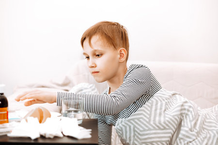 Sick sad little kid teen boy sits on bed at home and is going to take medication. Flu season. Healthcare conceptの写真素材