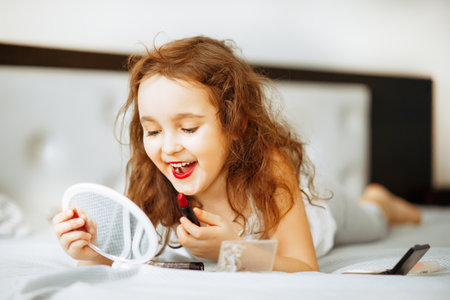 Kids makeup. Happy cute girl doing makeup lies on bed in the bedroom. Kid using lipstick looks in the mirror. Skin care concept. Cosmetics for childrenの写真素材