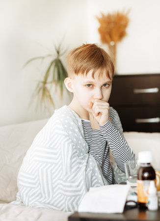 Sick little teen boy sits on bed at home and coughs. Asthma medicine. Flu season. Healthcare conceptの写真素材
