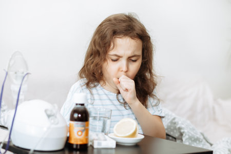Sick little kid girl sits on bed at home and coughs. Asthma medicine. Flu season. Healthcare conceptの写真素材