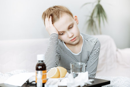 Sick sad little kid teen boy sits on bed at home and is going to take medication. Flu season. Healthcare conceptの写真素材