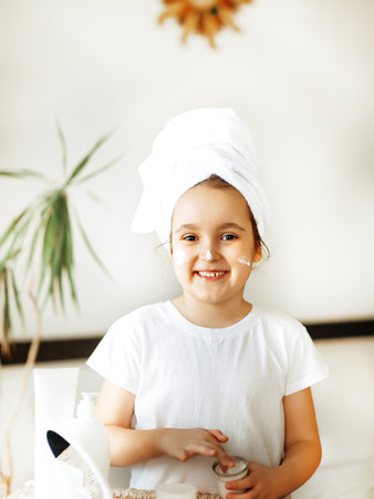 Funny little girl with towel on her head uses facial cream. Spa, healthcare, morning routine conceptの写真素材