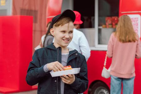 Cute teen boy kid eating hot dog nearby food track on a street. Food delivery or take away food conceptの写真素材