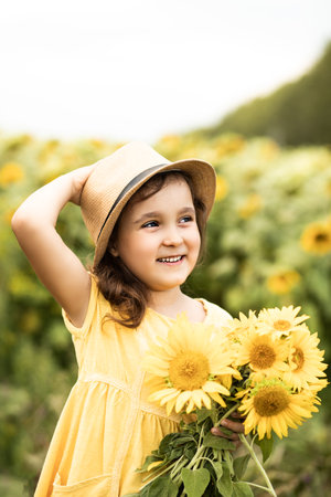 Smiling cute girl in a hat and yellow dress with bouquet of sunflowers on field. Summer holidayの写真素材