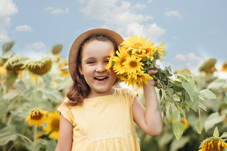 Smiling cute girl in a hat and yellow dress with bouquet of sunflowers on field. Summer holidayの写真素材