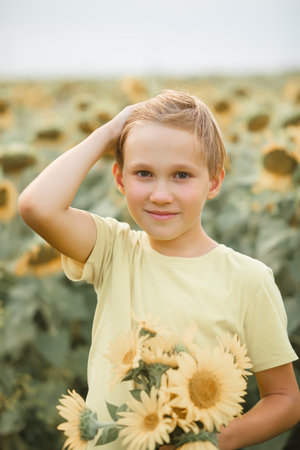 Smiling blonde cute kid boy teen standing on a field and holding bouquet of sunflowers. Summer holiday. Happy childhoodの写真素材