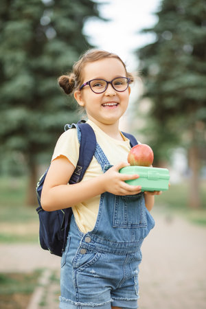 Cute little schoolgirl with backpack holding in a hands lunch box. Near by school outdoor. Food for kidsの写真素材