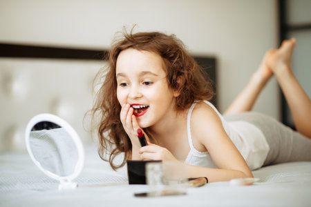 Kids makeup. Happy cute girl doing makeup lies on bed in the bedroom. Kid using lipstick looks in the mirror. Skin care concept. Cosmetics for childrenの写真素材