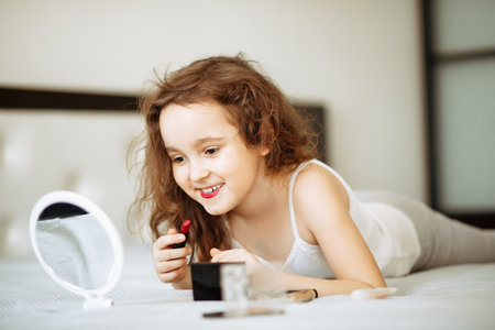 Kids makeup. Happy cute girl doing makeup lies on bed in the bedroom. Kid using lipstick looks in the mirror. Skin care concept. Cosmetics for childrenの写真素材