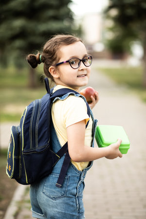 Cute little schoolgirl with backpack holding in a hands lunch box. Near by school outdoor. Food for kidsの写真素材